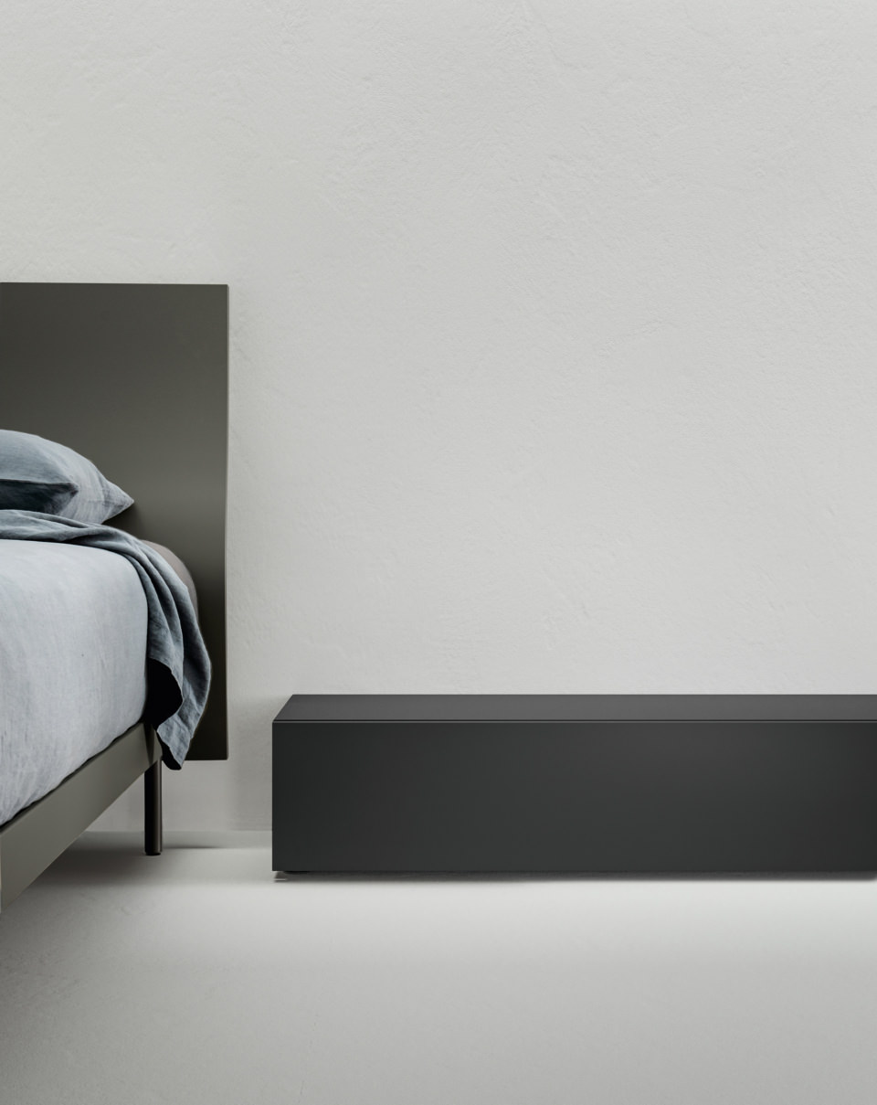 Una clara línea horizontal recoge la silueta de la cama y la desata en la pared contigua, creando una geometría moderna y empactante. 
El cajón está equipado con un mecanismo de retorno amortiguado para una experiencia aún más agradable. 
El acabado Nero Stout confiere autoridad a la pieza, que oculta el espacio de almacenamiento ideal para tu dormitorio. 